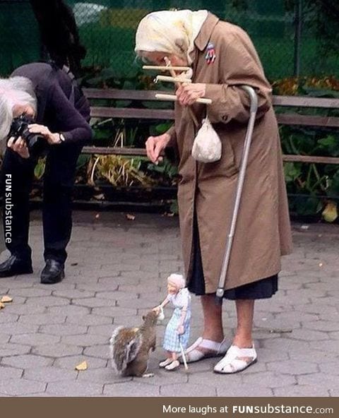 Old woman uses a marionette of herself to feed squirrels in the park